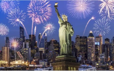 The Statue of Liberty stands tall in the foreground with a city skyline behind it. Fireworks burst in various colors in the night sky, creating a festive atmosphere. The lights from the buildings and fireworks illuminate the scene. 10 Cheap Travel Destinations In the United States