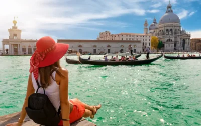 A woman in a red hat and skirt sits on the edge of a wooden platform, overlooking a canal in Venice. Gondolas with passengers float by on the turquoise water, while historic buildings and a domed church are visible in the background under a clear blue sky. Affordable Travel Destinations