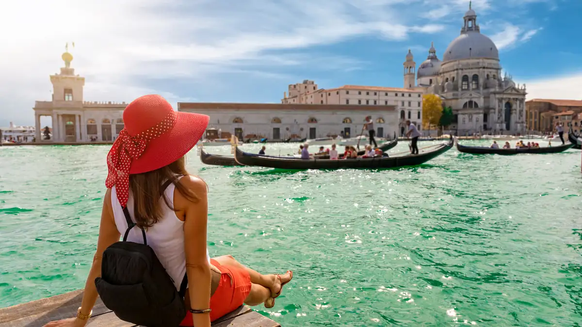 A woman in a red hat and skirt sits on the edge of a wooden platform, overlooking a canal in Venice. Gondolas with passengers float by on the turquoise water, while historic buildings and a domed church are visible in the background under a clear blue sky. Affordable Travel Destinations