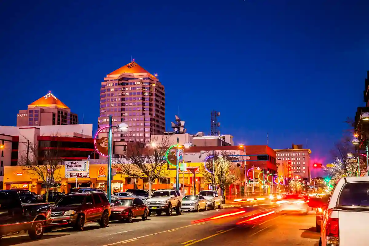 A vibrant city street scene at night, with cars parked along the sides and others driving by, leaving light trails. Tall buildings with illuminated tops stand in the background under a dark blue sky. Neon street decorations adorn the sidewalks.
