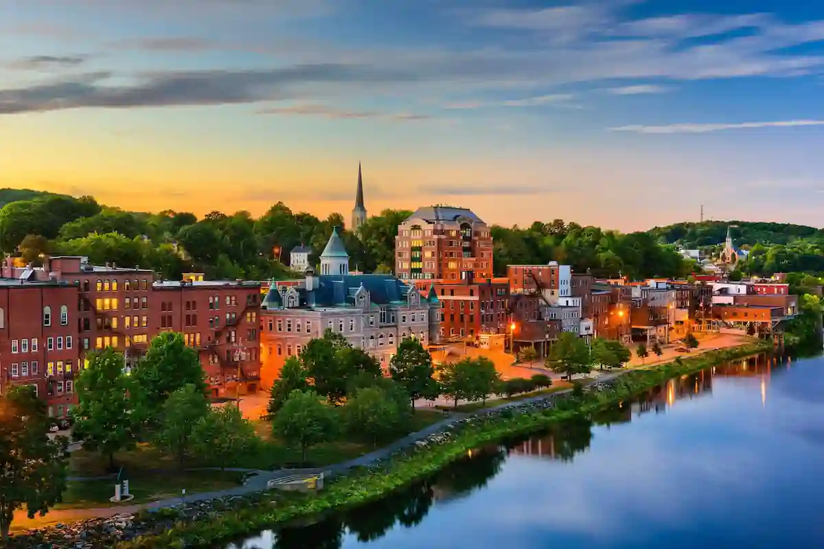 Scenic view of a charming town alongside a serene river at sunset. Historic buildings with red brick facades and a prominent church steeple are illuminated by street lights, surrounded by lush greenery. A calm sky with warm hues adds to the picturesque setting.
