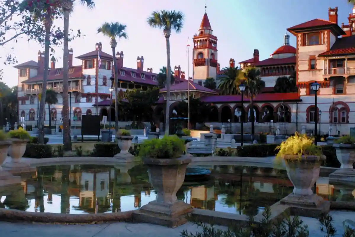 A historic building with red-tiled roofs and arches is reflected in a serene pond surrounded by lush greenery and palm trees. The sky is clear, enhancing the picturesque and tranquil atmosphere of the scene.
