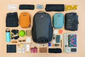 A neatly arranged travel packing layout includes a black backpack, two smaller packing cubes (one orange, one teal), a rolled jacket, various clothing items, toiletries, a water bottle, a wallet, sunglasses, a phone, and other travel essentials on a beige background. Travel Essentials for Anxiety