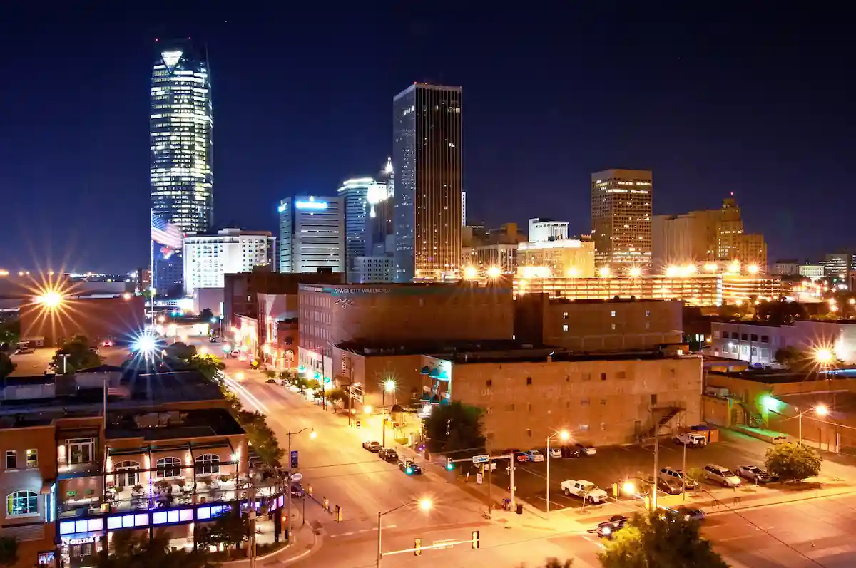 A vibrant cityscape at night featuring tall, illuminated skyscrapers and various buildings along well-lit streets. The tallest building, glowing brightly, stands out against the dark sky. The streets below are busy with light trails from moving vehicles.
