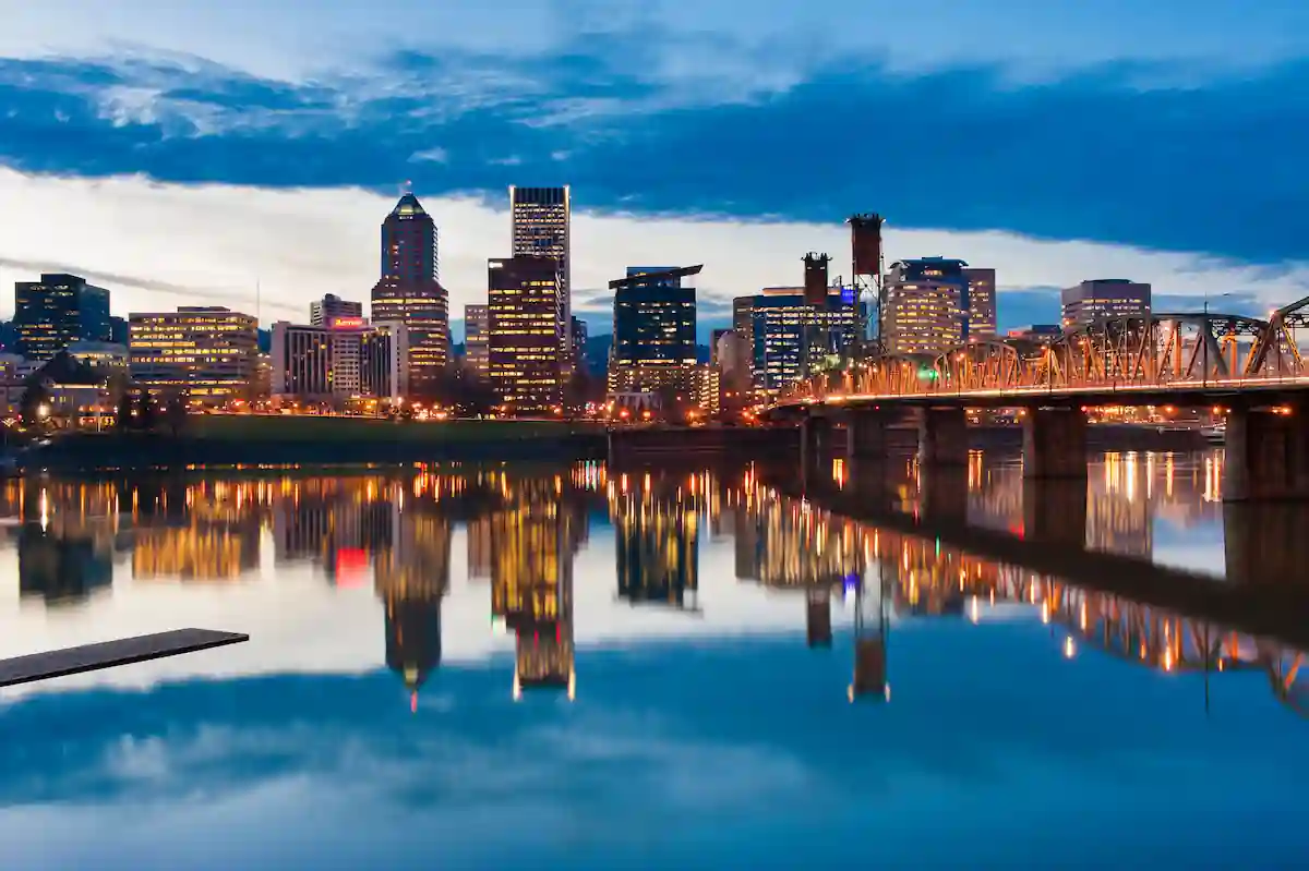 A cityscape at dusk featuring illuminated skyscrapers and buildings reflecting on the calm water of a river. A bridge spans the river, connecting both sides of the city, under a cloud-laden sky tinged with the glow of the setting sun.
