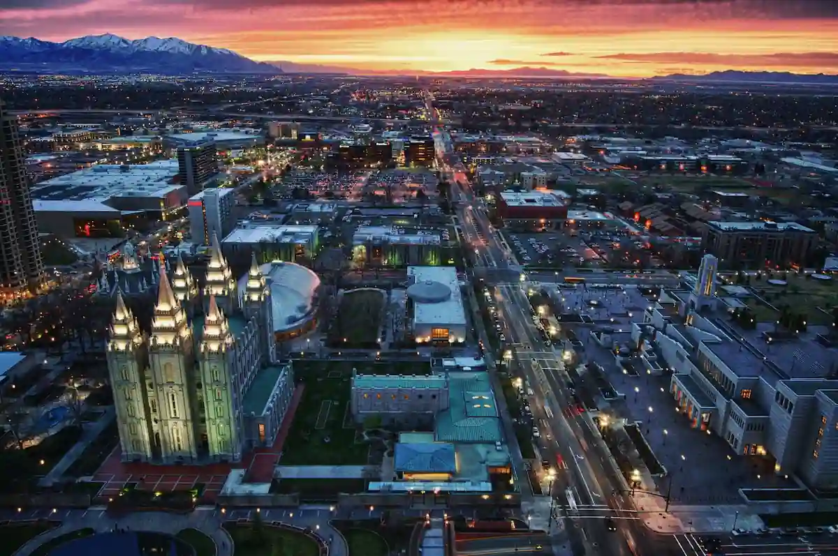 Aerial view of Salt Lake City during sunset. Prominently visible is the Salt Lake Temple with its illuminated spires. Surrounding buildings and streets are lit up as night falls, and distant mountains are outlined against the colorful sky.
