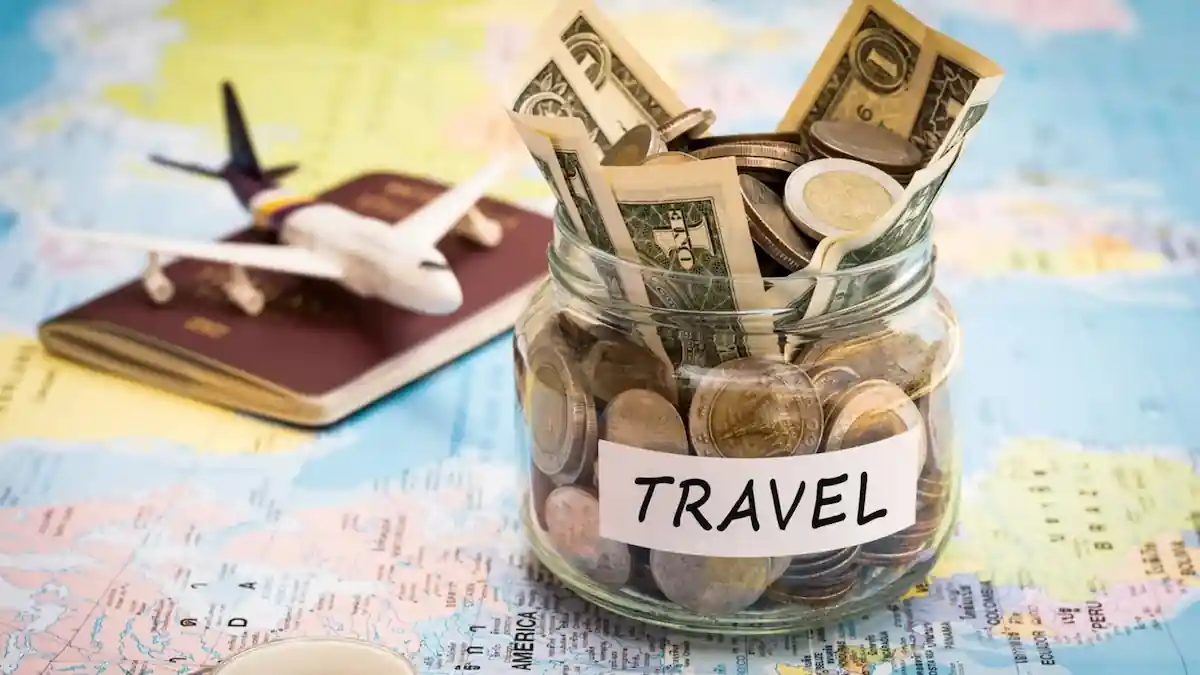 A glass jar labeled "TRAVEL" filled with various currencies including banknotes and coins is placed on a world map. In the background, there is a toy airplane and a passport.
