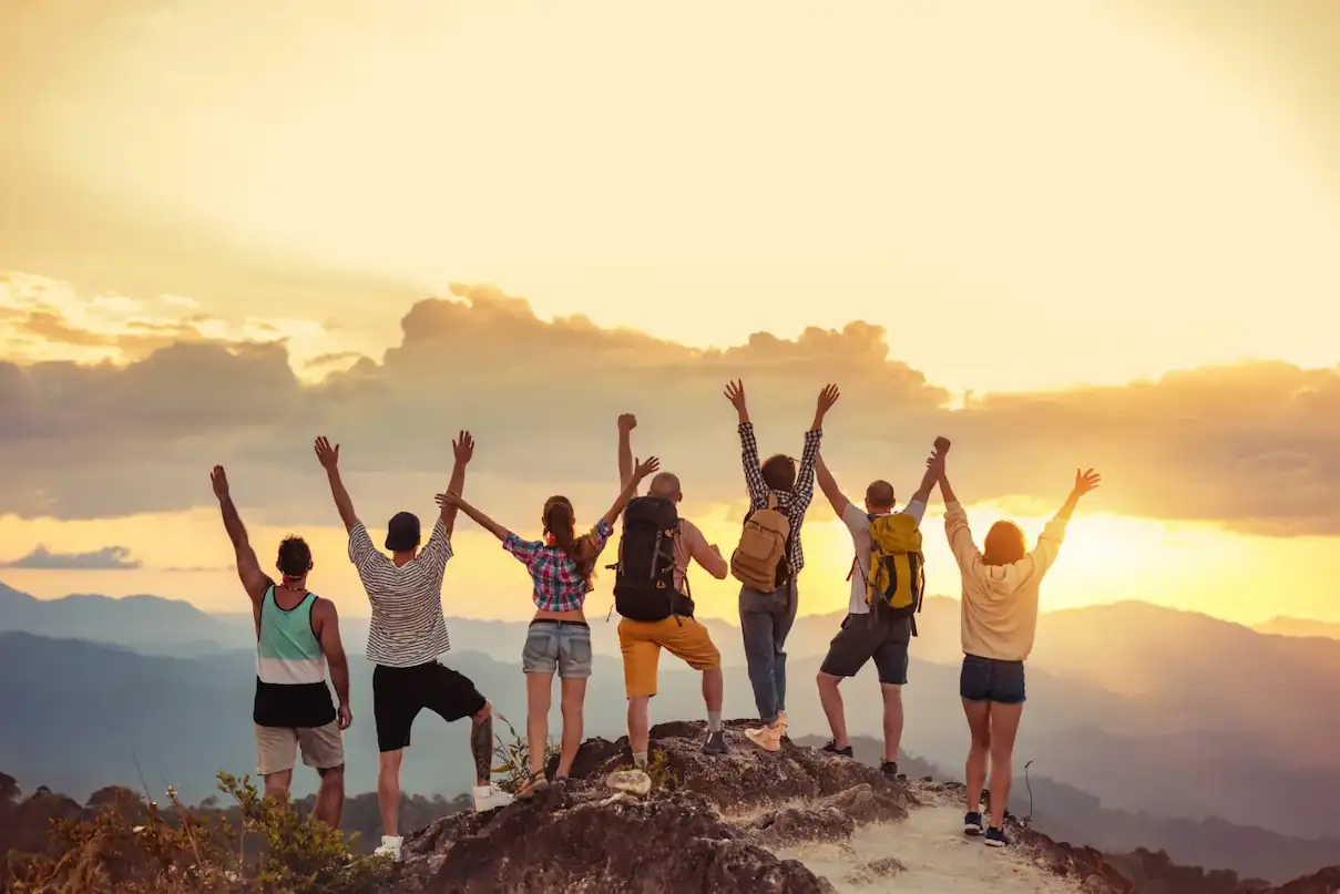 Six people stand on a rocky hilltop at sunset, facing away from the camera and holding their arms up in celebration. They are dressed in casual outdoor clothing, some with backpacks, surrounded by a scenic landscape of distant mountains and a dramatic sky. Solo Travel Groups