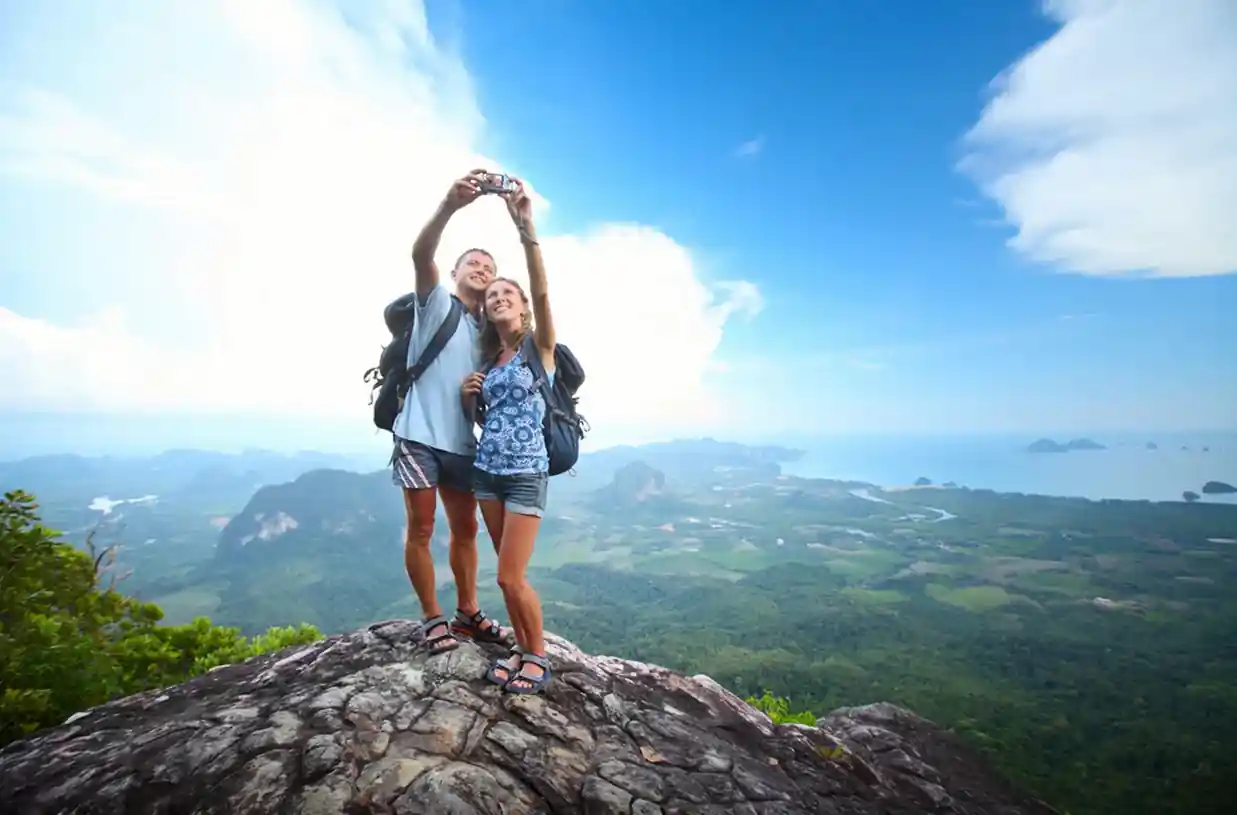 A couple stands on a rocky cliff taking a selfie against a backdrop of lush green valleys and distant mountains. They are wearing backpacks and casual clothing, and the sky is bright and partly cloudy.
 Solo Travel Group and Packages