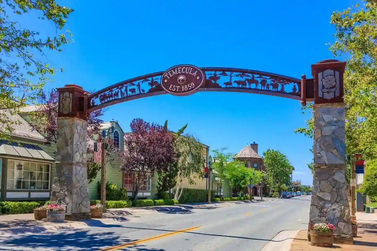 A street scene showcasing an arched sign reading "TEMECULA EST. 1859" supported by two stone pillars. The sign features decorative elements and silhouettes of horses. Trees and traditional buildings line the street under a bright blue sky.
