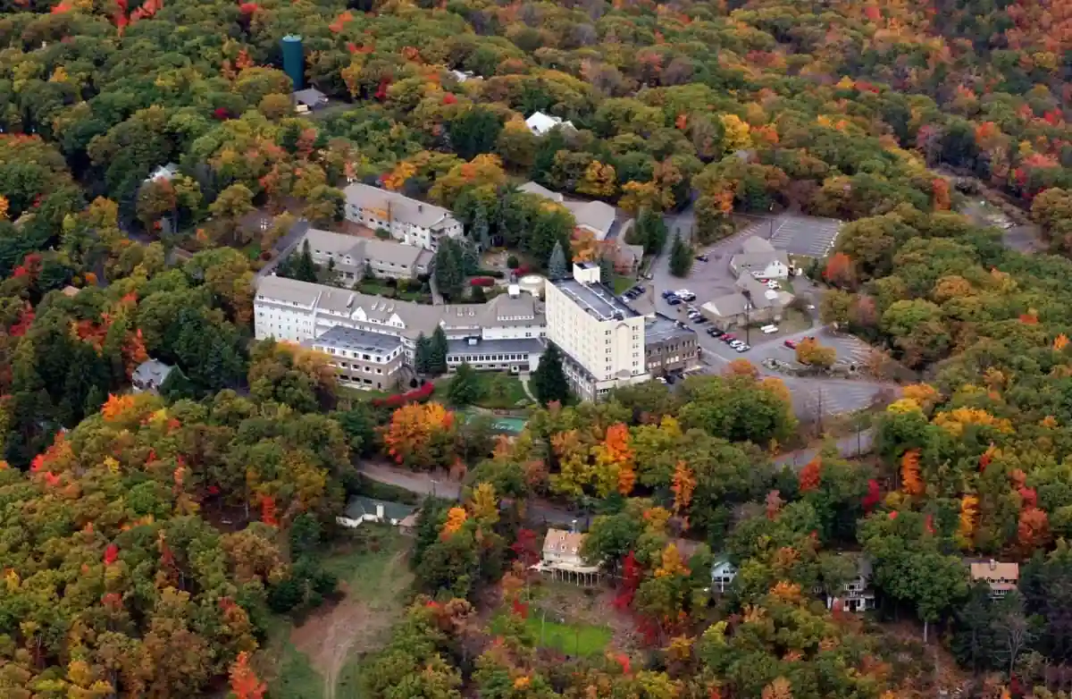 Aerial view of a large building complex surrounded by dense trees displaying vibrant fall foliage in shades of green, yellow, orange, and red. The complex includes several buildings, parking areas, and a few small structures scattered around the wooded area.
