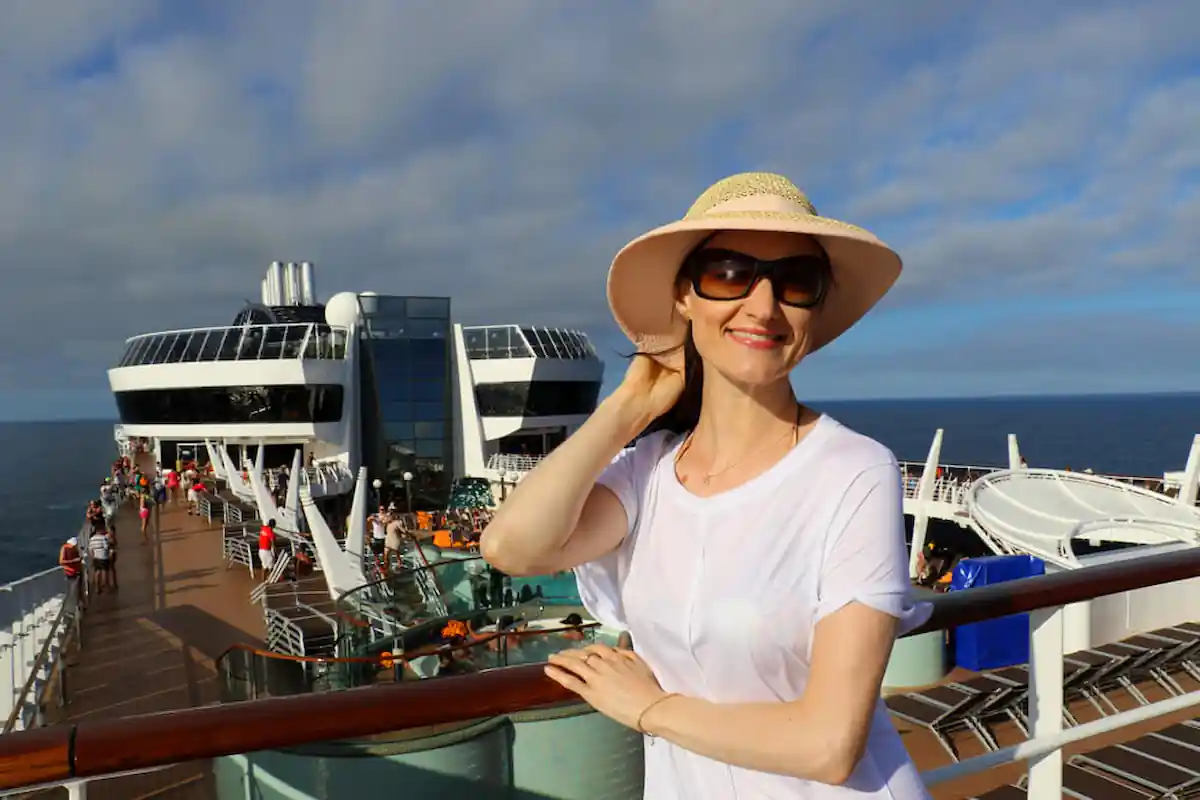 A woman wearing a sun hat and sunglasses is smiling while leaning on a railing on a cruise ship. The ship's upper deck is busy with other passengers, and the ocean and a partly cloudy sky are visible in the background.
Travel Groups for Singles Over 40 to 50 singles 