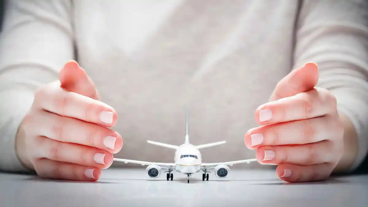 A small model airplane is placed on a flat surface, surrounded by two hands gently cupping around it, suggesting protection or care. The blurred background implies a focus on the plane and hands.
