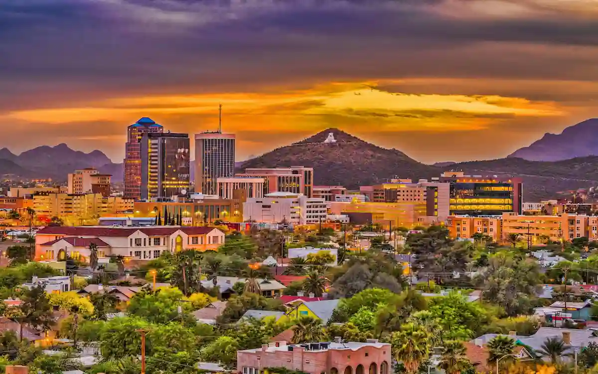 A vibrant cityscape of Tucson, Arizona, at sunset. The sky displays dramatic hues of orange, yellow, and purple. Modern buildings are seen in the foreground, with the "A" Mountain and surrounding hills in the background.
