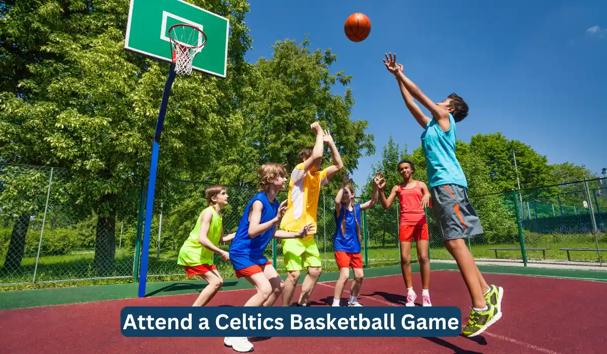 A group of kids engaged in a lively outdoor basketball game, jumping and reaching for the ball near a green basketball hoop. The background features lush green trees under a clear blue sky. A text overlay at the bottom reads "Attend a Celtics Basketball Game.
