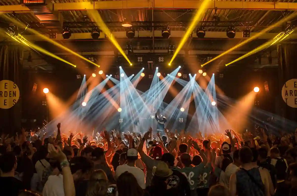 A large crowd enjoys a concert in an indoor venue. The stage is lit up with vibrant lights in various colors, including yellow, orange, and white. The audience has their hands up, engaging with the performance. The atmosphere is energetic and lively.
