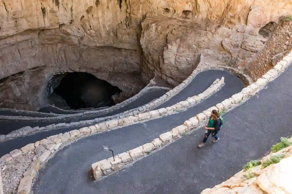 A large, natural cave opening with a spiral walkway leading down. A woman is walking down the path, exploring the cave. The walls of the cave are made of rock and have a natural, rough texture.