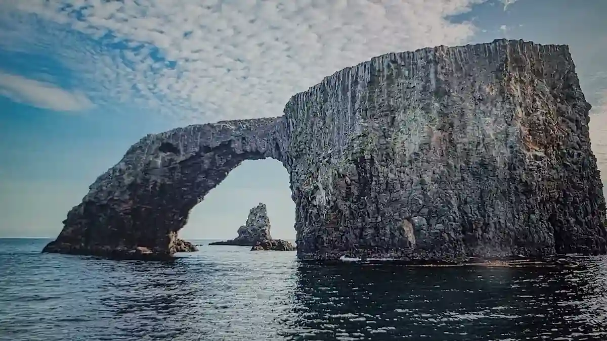 A large, natural rock arch rises from the ocean, creating a dramatic frame against a backdrop of blue sky and white clouds. Smaller rocky outcroppings dot the water around the arch.