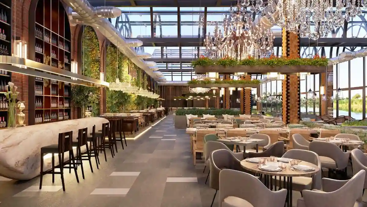 A spacious, modern restaurant with high ceilings and large windows letting in natural light. The interior features contemporary chandeliers, a mix of upholstered chairs and wooden tables, a long bar with stools, and abundant greenery throughout the space.
