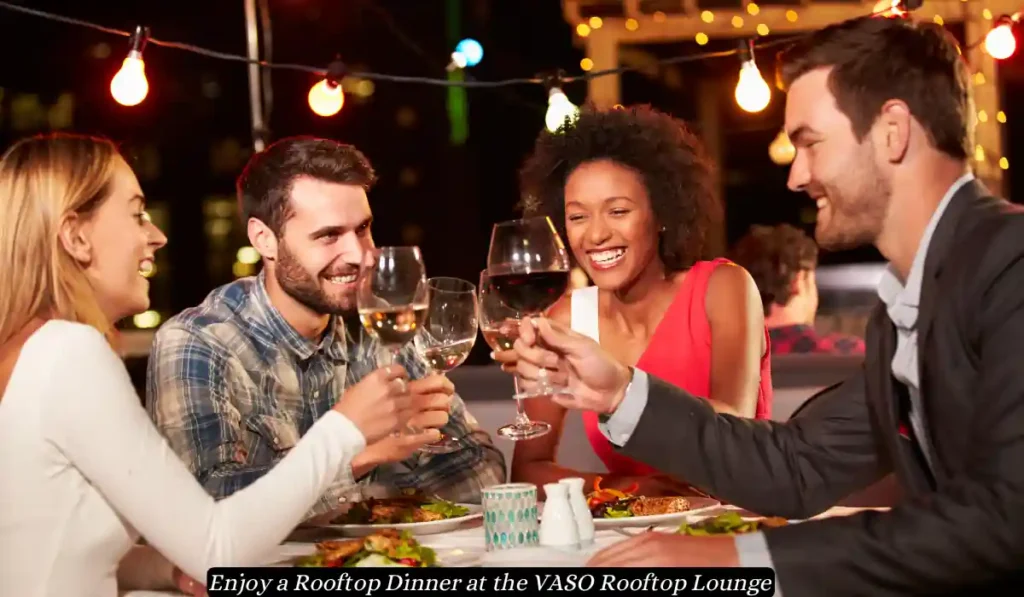 A group of four friends, two women and two men, sit around a table clinking wine glasses while smiling and enjoying dinner at a rooftop lounge adorned with string lights. The text overlay reads, “Enjoy a Rooftop Dinner at the VASO Rooftop Lounge.”.
