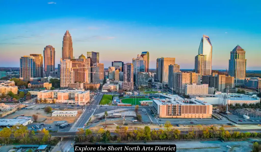 Aerial view of a city's downtown area during the early evening. Numerous tall buildings and skyscrapers are visible under a clear sky. The scene is bustling with various structures, with green spaces and roads in the foreground. Text at the bottom reads: "Explore the Short North Arts District.
