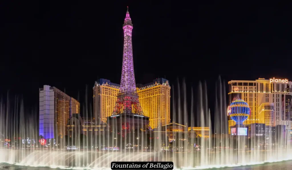 A nighttime view of the Las Vegas Strip featuring the Fountains of Bellagio. The lights of the Paris Las Vegas Hotel, including the Eiffel Tower replica, are brightly illuminated in the background, along with other iconic casino and hotel signs.
