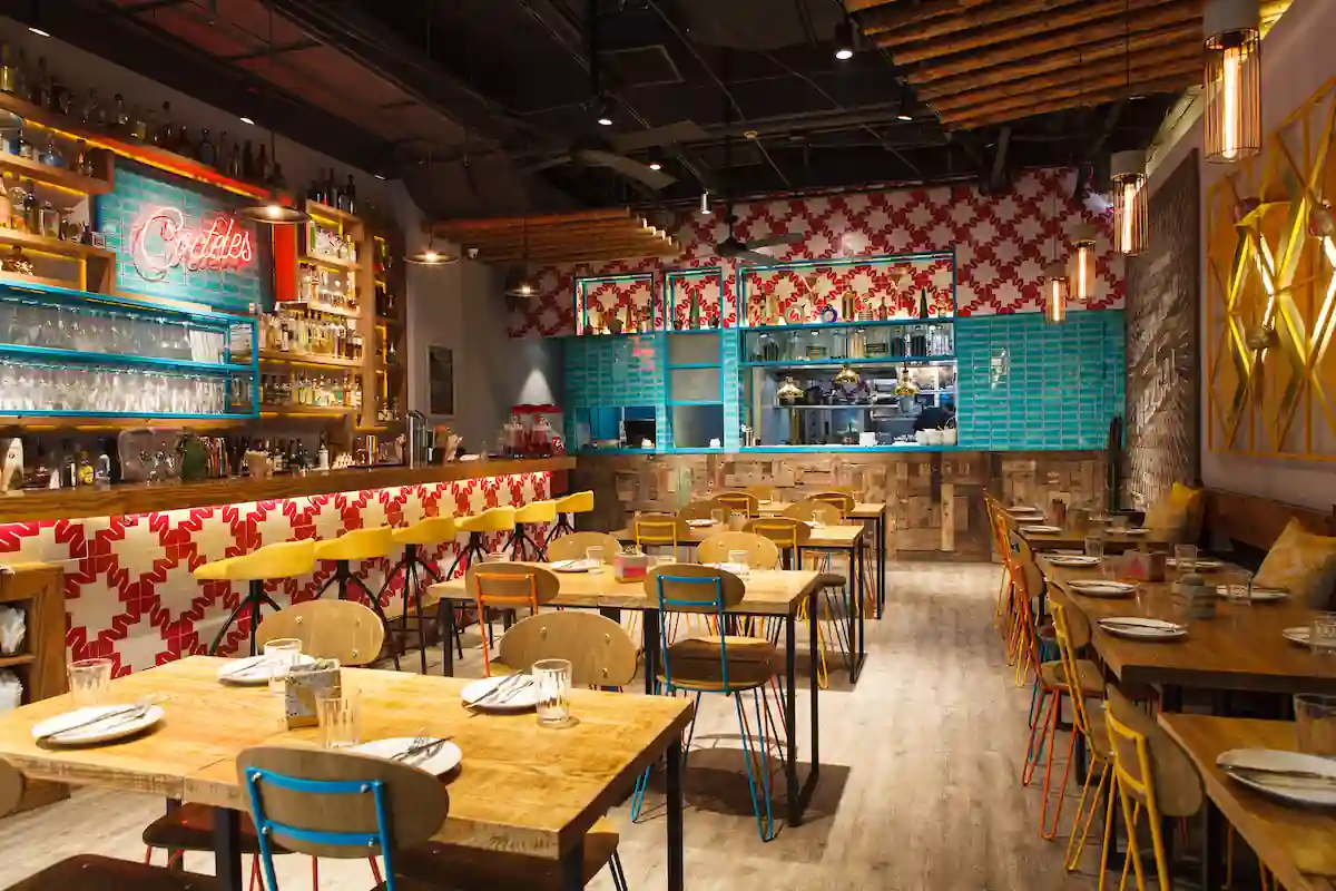 A modern restaurant interior with wooden tables and chairs, a bar area with the neon sign "Cocktails," and a partially open kitchen in the background. The walls are decorated with geometric red and white patterns, and the overall atmosphere is warm and inviting. .
