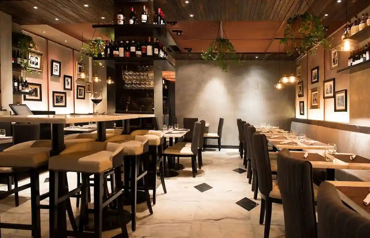 A cozy, modern restaurant with a warm atmosphere. The space includes tables with cushioned high chairs and standard chairs, ambient lighting, and wall art. Wine bottles and plants are displayed on shelves, adding to the inviting decor. Marble flooring enhances the elegance.
