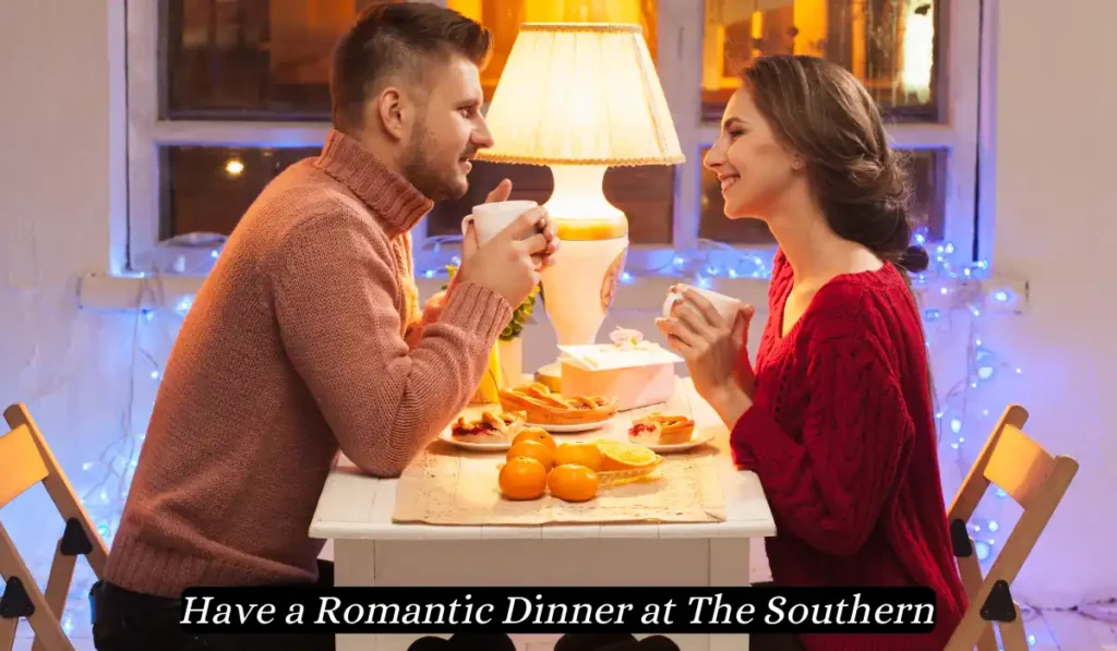 A couple sits across from each other at a small, white table, enjoying a cozy dinner. The table is adorned with an assortment of food and lit by a warm lamp, creating an intimate atmosphere. The image caption reads, "Have a Romantic Dinner at The Southern.
