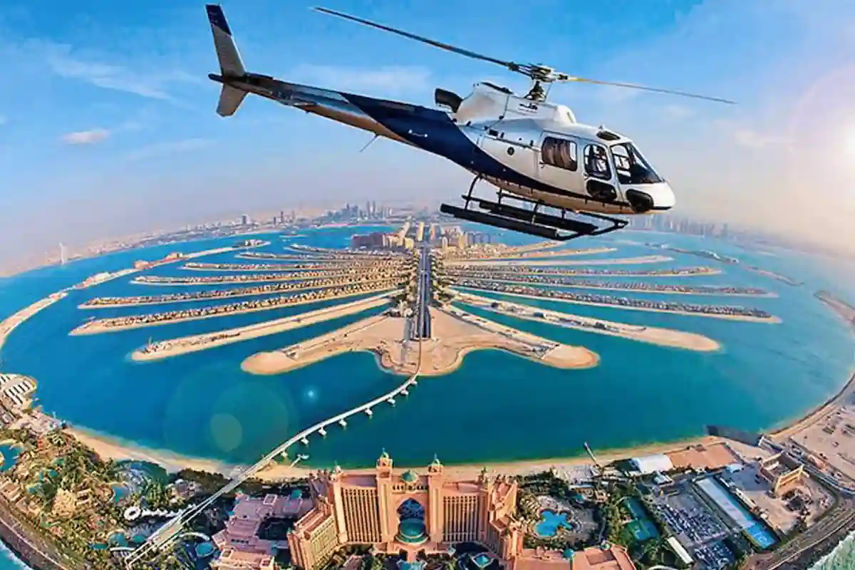 A helicopter flies over the iconic Palm Jumeirah in Dubai, United Arab Emirates. The aerial view showcases the man-made palm tree-shaped island, surrounded by blue waters, with the Atlantis Hotel visible in the foreground. The city skyline is in the distance.
 