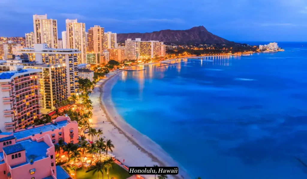 Aerial view of Waikiki Beach in Honolulu, Hawaii, at dusk. The city skyline is lit up with numerous buildings, while the calm waters of the Pacific Ocean reflect the lights. The iconic Diamond Head crater is visible in the background.
