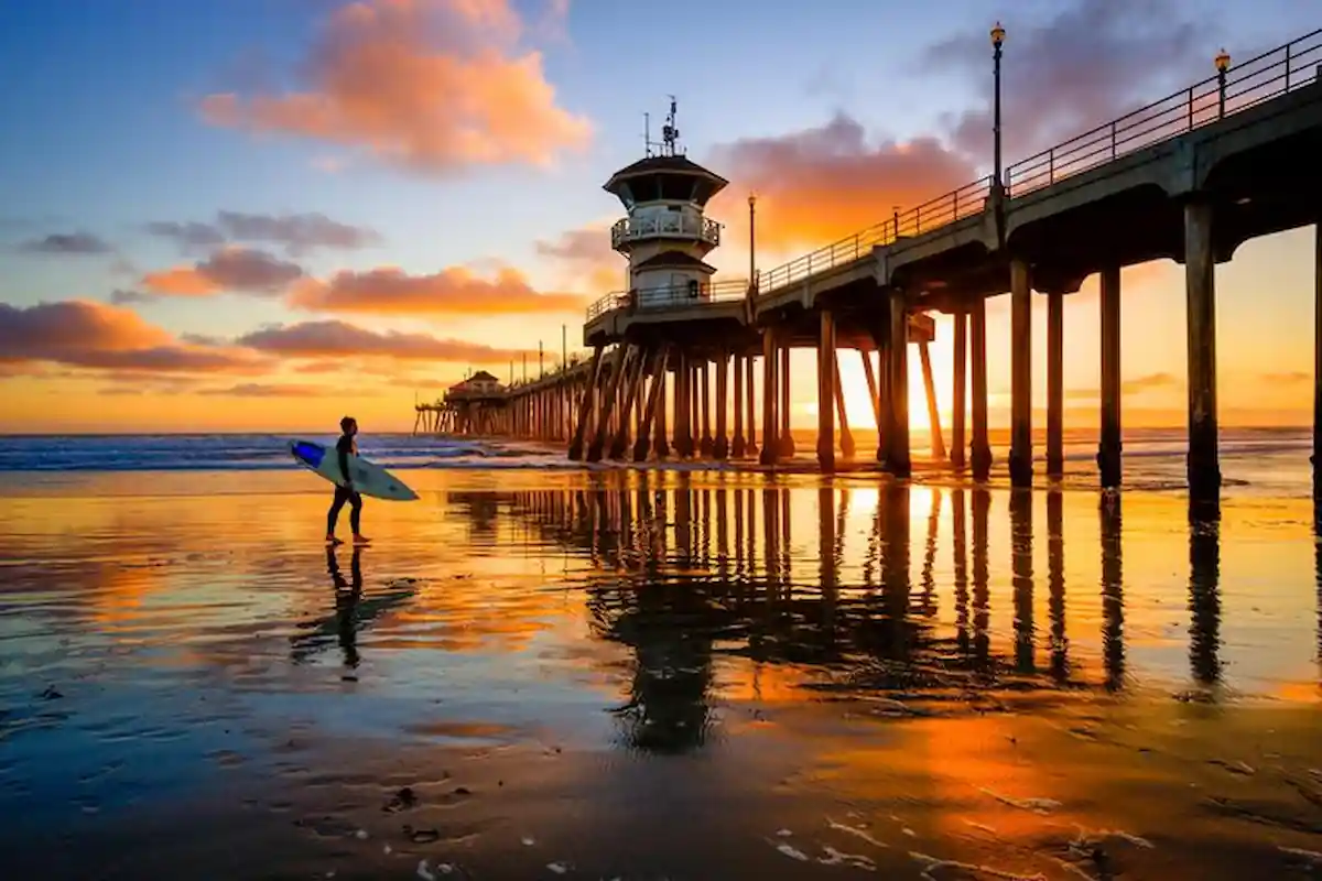 A surfer walks along a beach at sunset. A long pier with a tower extends into the ocean. The sky is filled with colorful clouds, and the ocean reflects the golden light.