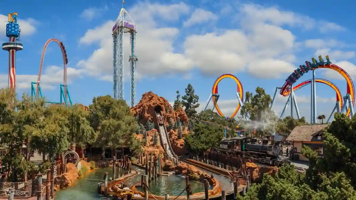 This image shows a vibrant amusement park with various roller coasters, a tall tower ride, and a water ride. The park is surrounded by trees and has a Western theme with a rock formation and a steam train.