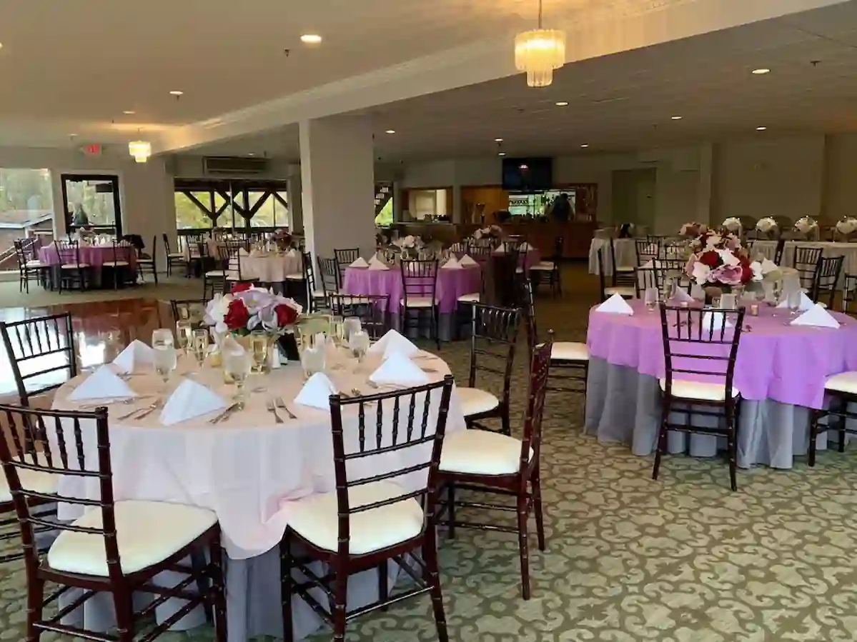 A spacious banquet hall with round tables elegantly set for an event. Tables feature white tablecloths, pink overlays, floral centerpieces, neatly folded white napkins, and wooden chairs with cushioned seats. The room is softly lit by chandeliers and natural light.
