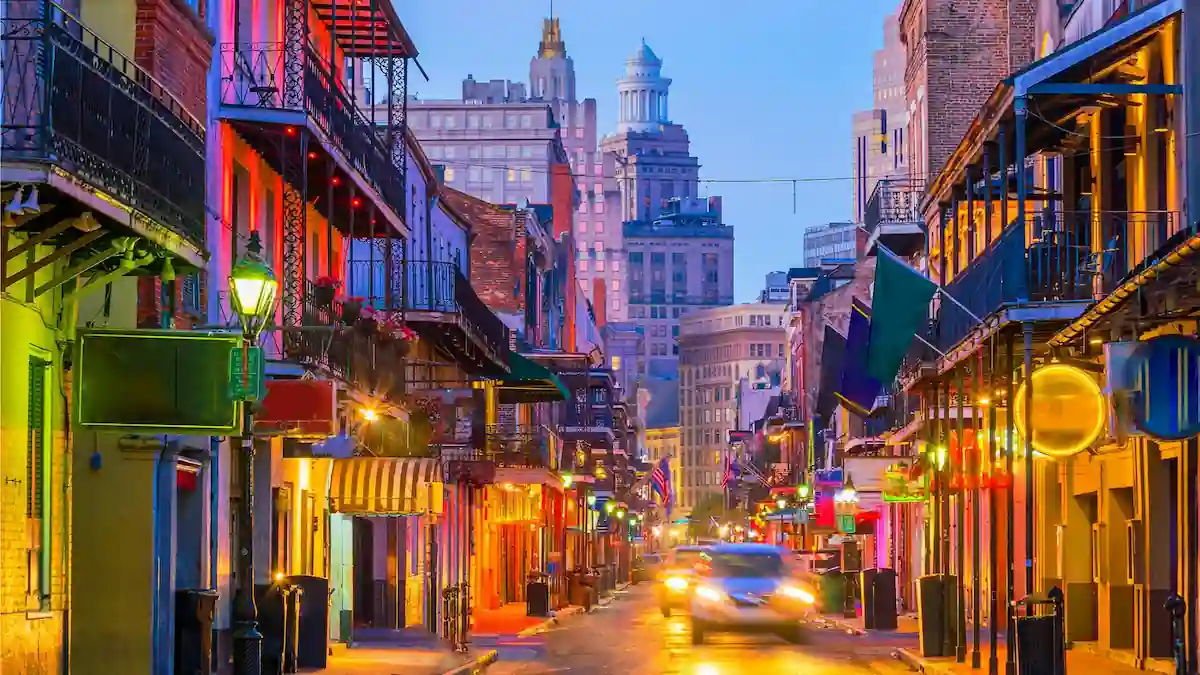 A vibrant street scene in the French Quarter of New Orleans at twilight. The street is lined with colorful buildings, neon lights, and balconies. A few cars blur past, emphasizing the lively atmosphere. In the background, taller buildings are illuminated.
