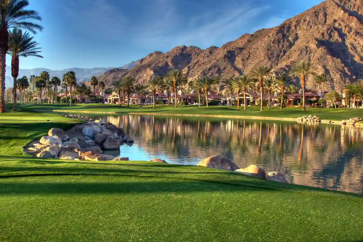 A scenic golf course surrounded by palm trees and mountains. A large lake reflects the blue sky and mountains. The green grass is lush and well-maintained. Houses can be seen in the distance.