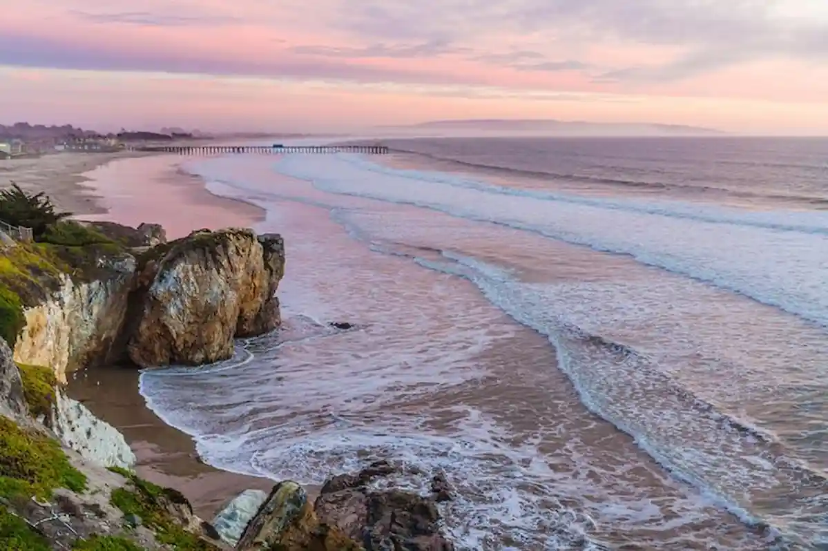 A scenic coastal landscape at sunset. The sky is ablaze with pink and purple hues, reflecting on the calm ocean waves. A long pier stretches out into the water, and a rocky cliff overlooks the sandy beach. The overall atmosphere is peaceful and serene.