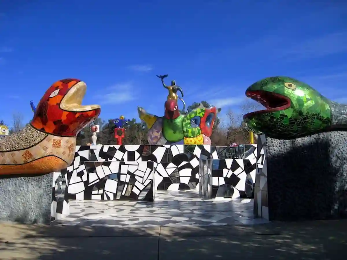 A vibrant, whimsical outdoor sculpture installation featuring two large, colorful mosaic serpents facing each other. A central mosaic structure with a humanoid figure stands between them. The background is a clear blue sky. This appears to be the Queen Califia's Magical Circle in Balboa Park, San Diego, California.