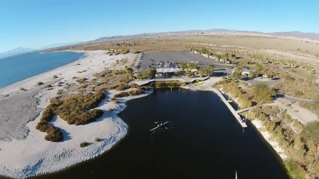 An aerial view of a coastal area with a large body of water. There is a sandy beach on one side and a man-made pond on the other. Buildings and parking lots are visible in the background. Mountains can be seen in the distance.