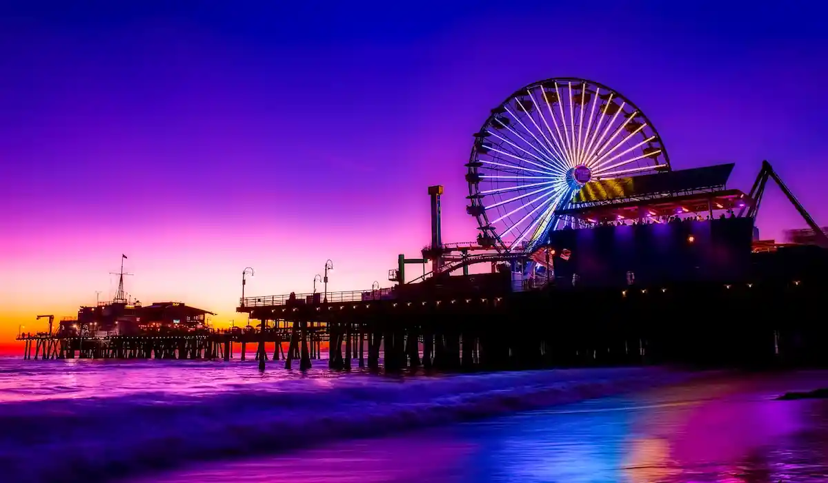 A brightly lit Ferris wheel on a pier at sunset. The sky is a vibrant purple and blue, and the ocean waves are crashing against the shore. There are other buildings and attractions visible on the pier.