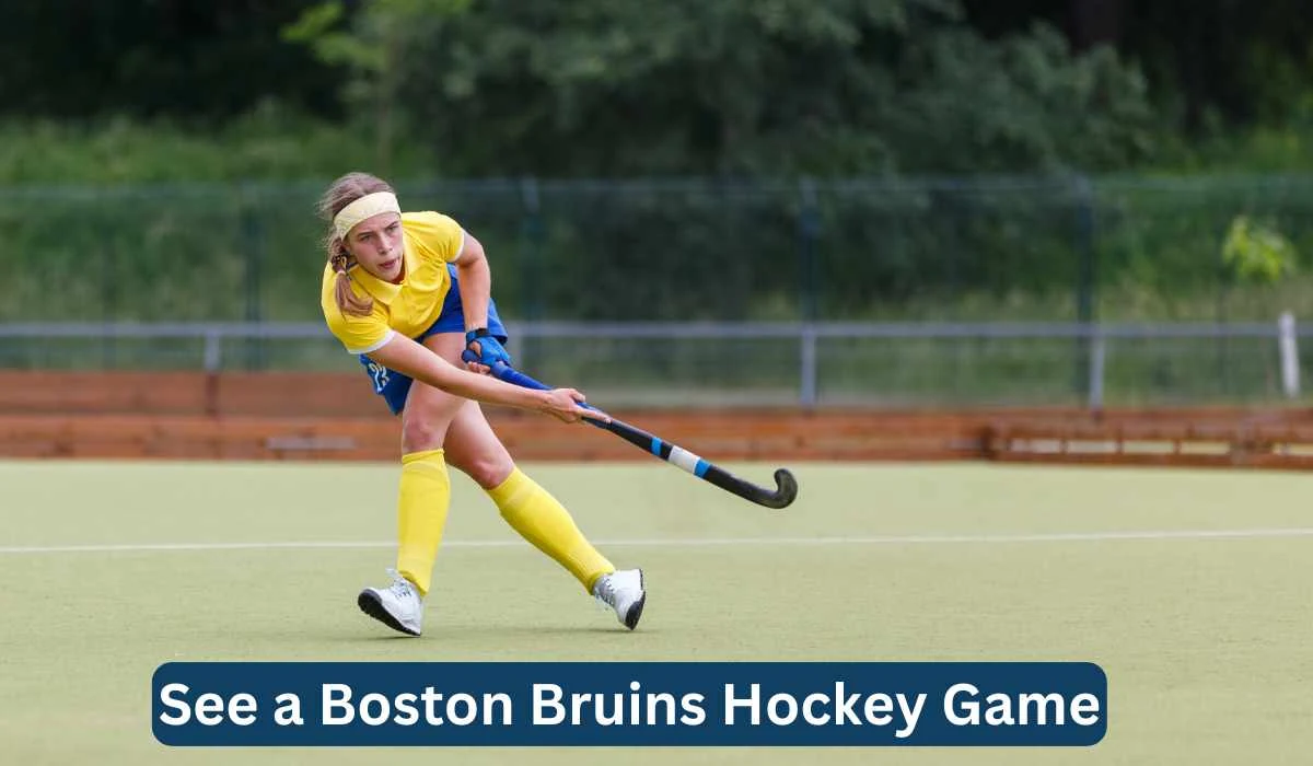 A young athlete in a yellow jersey and shorts plays field hockey on a green field, actively swinging a hockey stick. The surrounding area is blurred, indicating movement. The image has a caption reading, "See a Boston Bruins Hockey Game.
