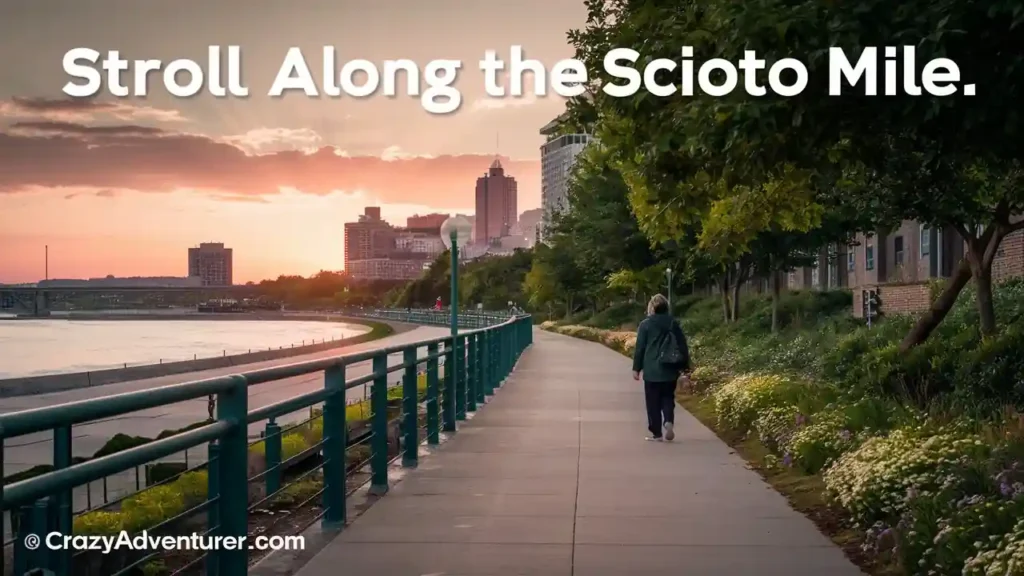 A person walks along a riverside path lined with trees and flowers at sunset. Tall buildings are visible in the background. The text "Stroll Along the Scioto Mile" is displayed at the top. The bottom left corner credits CrazyAdventurer.com.
