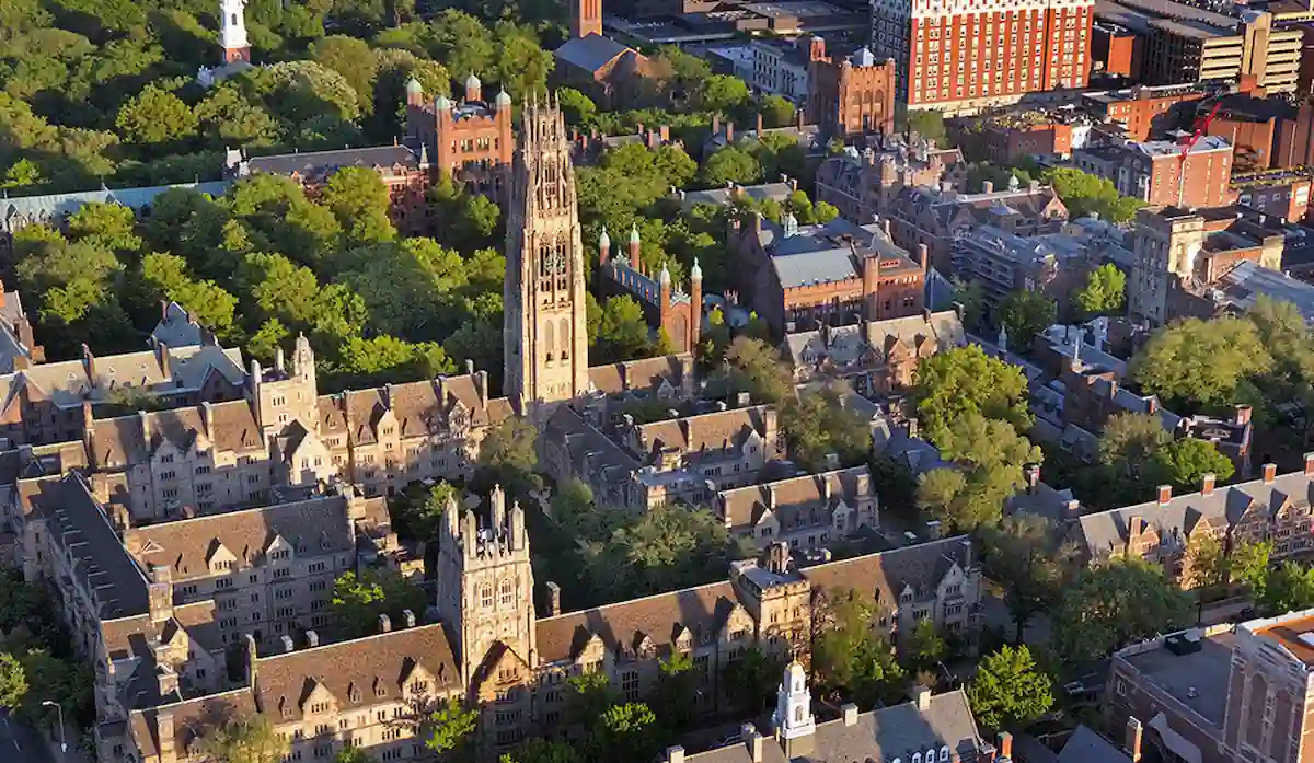 Aerial view of a historic university campus with numerous Gothic-style buildings, prominent spires, and lush green trees. A tall bell tower stands at the center, surrounded by various academic buildings and residential areas.
