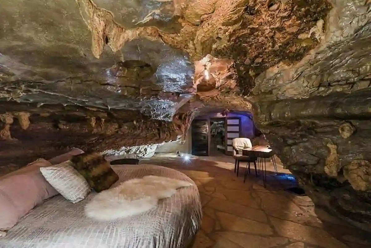 A unique bedroom carved into a natural cave. The room features a large, circular bed, stone flooring, and exposed rock walls adorned with stalactites. A small sitting area with a desk and chairs is visible in the background. The overall atmosphere is cozy and rustic.