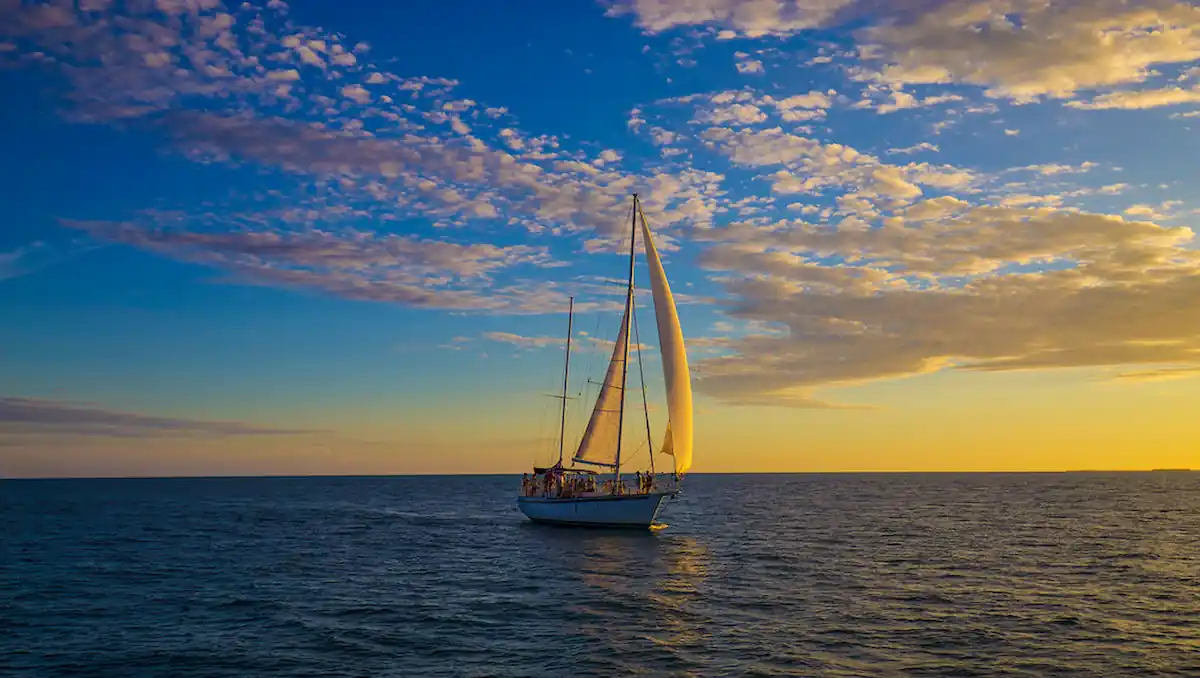 A sailboat with white sails glides on a calm sea during sunset. The sky is vibrant with hues of blue, yellow, and orange, and scattered with fluffy clouds. The boat's silhouette is prominently visible against the colorful sky.
