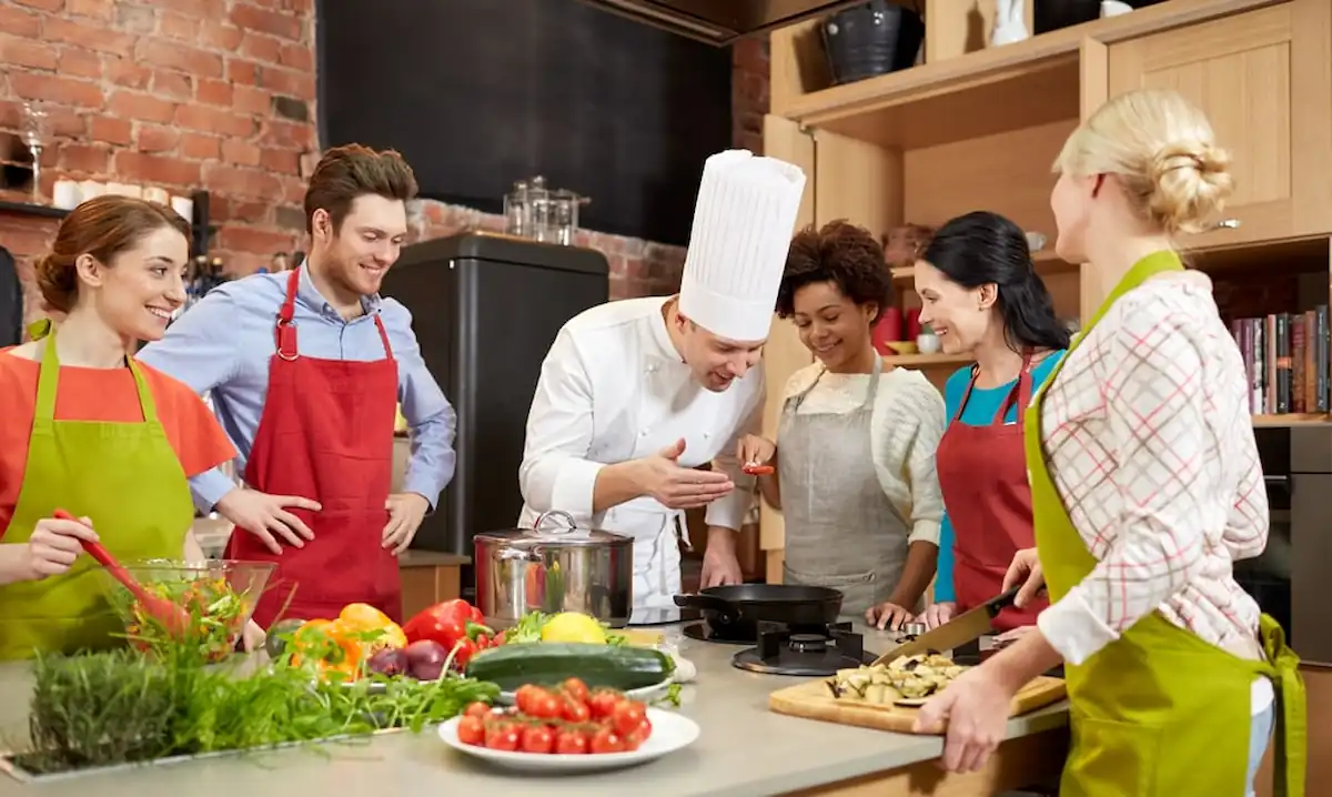 A chef in a white uniform and hat is demonstrating cooking techniques to a group of five adults in a kitchen. They are all wearing colorful aprons and look engaged. Various fresh vegetables are on the counter, and a woman is chopping vegetables while others watch.
