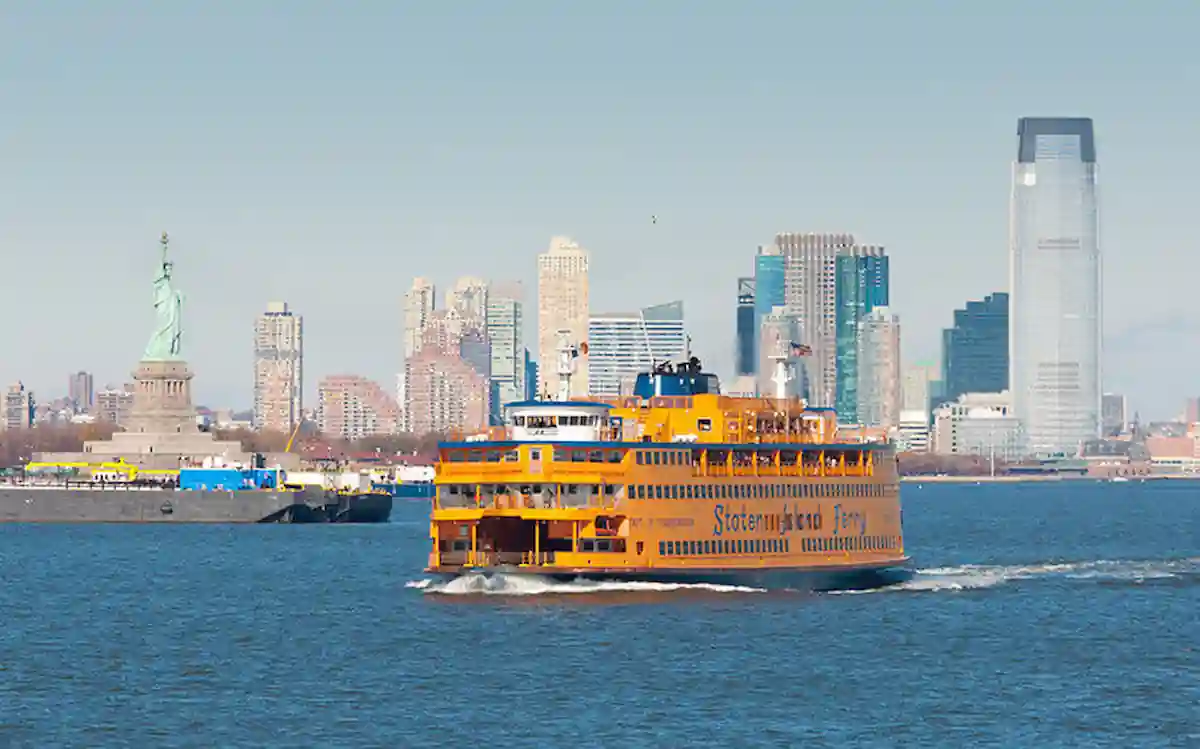 A bright orange Staten Island Ferry glides across the water with the Statue of Liberty on the left and the New York City skyline with tall buildings in the background under a clear blue sky.
