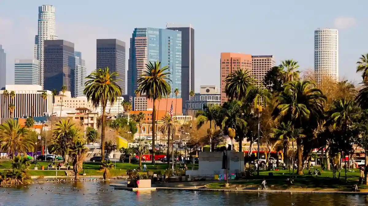 A sunny day in a park with lush palm trees near a calm body of water, set against the backdrop of modern skyscrapers in downtown Los Angeles. The scene captures the blend of nature and urban life, with people enjoying the park and the iconic skyline towering in the distance.