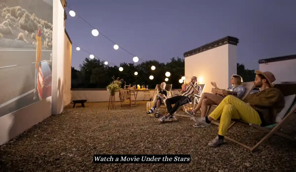 A group of people seated on outdoor lounge chairs watch a movie projected onto a large wall at night. The scene is lit by string lights hanging above. A caption reads, "Watch a Movie Under the Stars." The setting has a cozy, rustic ambiance with a relaxed vibe.
