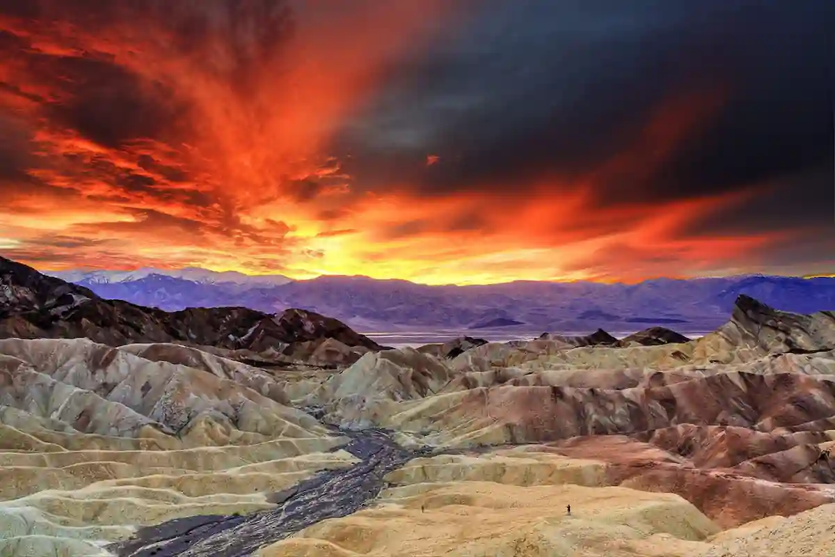 A breathtaking sunset over Death Valley National Park. The sky is ablaze with fiery orange and red hues, casting dramatic shadows on the colorful, eroded hills below. A small stream winds its way through the valley, and tiny figures of hikers can be seen on the distant hills.